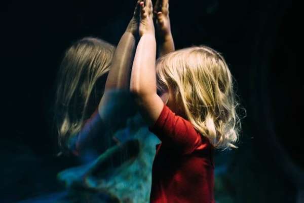 Girl leaning on glass fish tank raising her two hands, creating a triangle with her reflection.