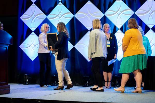 Three U.S. WIN members walk across the stage to receive chapter awards while the U.S. WIN Awards & Recognition Committee shakes their hand. The picture shows Maryanne Stasko [left] and Lisa Zurawski [right] smiling at the Award WINners.